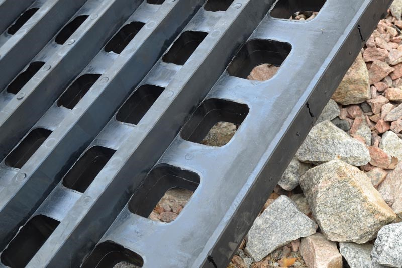 Svarta plattor med hål, liggande på grus och stenar.